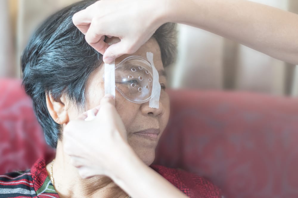 Caregiver gently applying a protective eye shield on an elderly woman’s eye post-surgery, emphasizing recovery and post-op eye protection.
