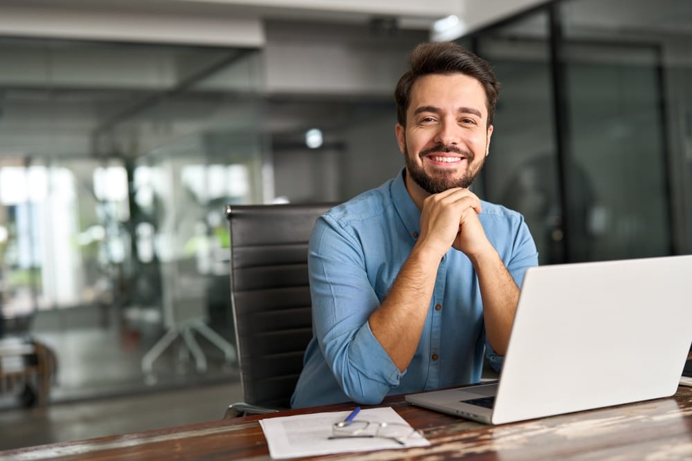 A cheerful man in a blue shirt sitting at a desk with a laptop and paperwork in a glass-walled office, representing modern work culture - Smart Technology