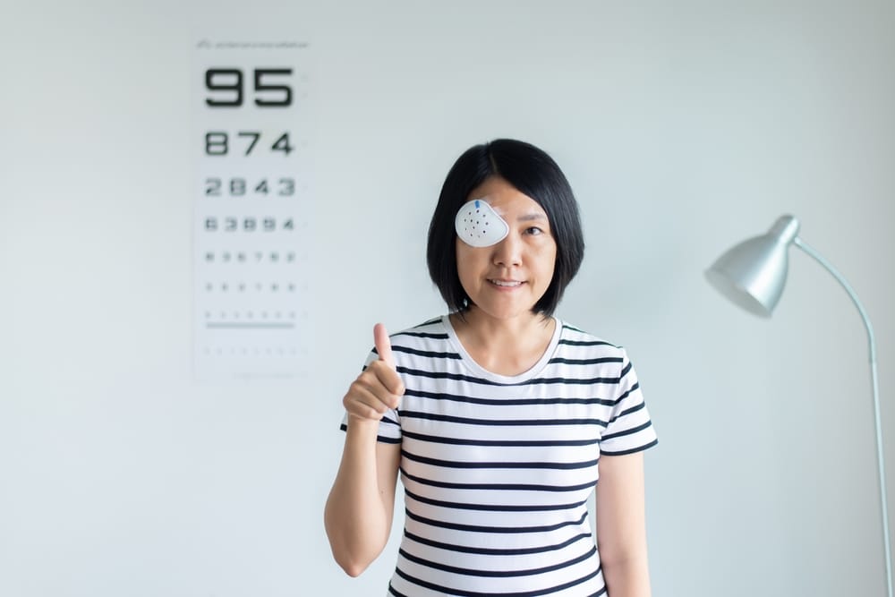 Woman undergoing a vision test with an eye occluder in front of a Snellen chart, used to assess visual acuity in an eye clinic setting.