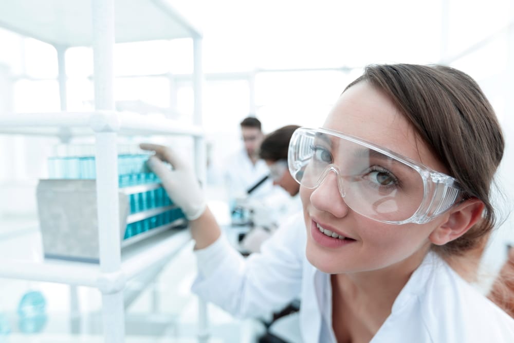 Smiling female lab technician wearing clear safety goggles while handling lab samples, showcasing proper eye protection in a research setting. 