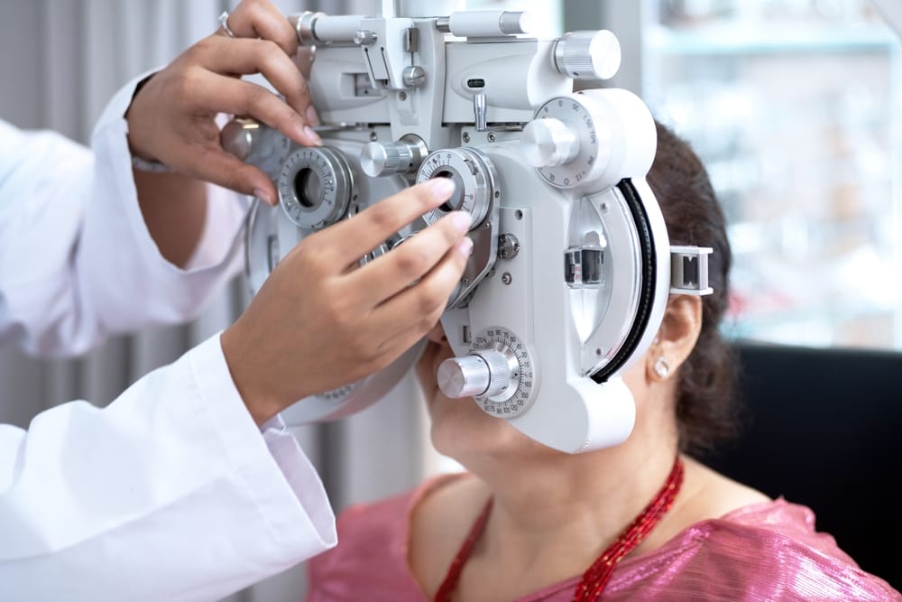 Eye doctor performing a refraction test using a phoropter to determine accurate eyeglass prescription for a female patient during her eye exam.