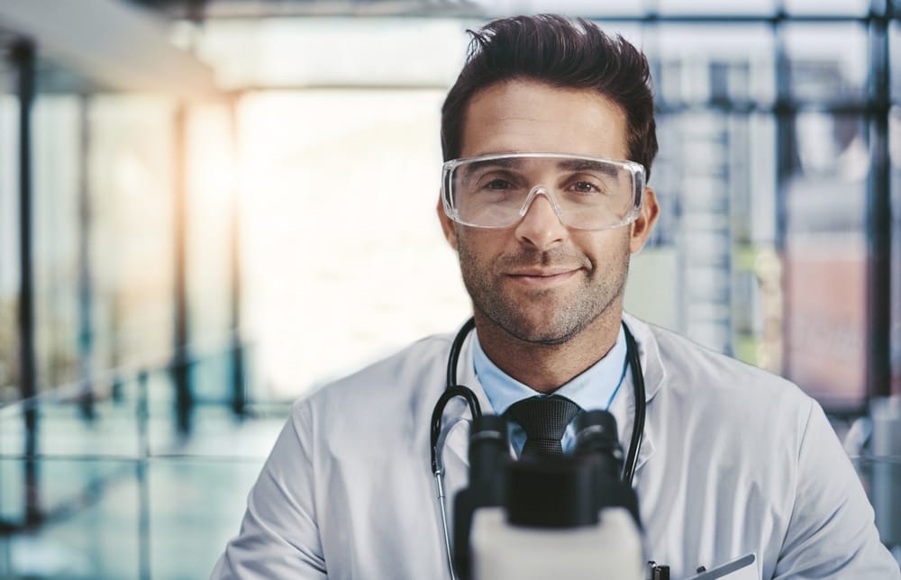 A confident male doctor in safety glasses and a stethoscope standing behind a microscope in a high-tech medical facility - Smart Technology