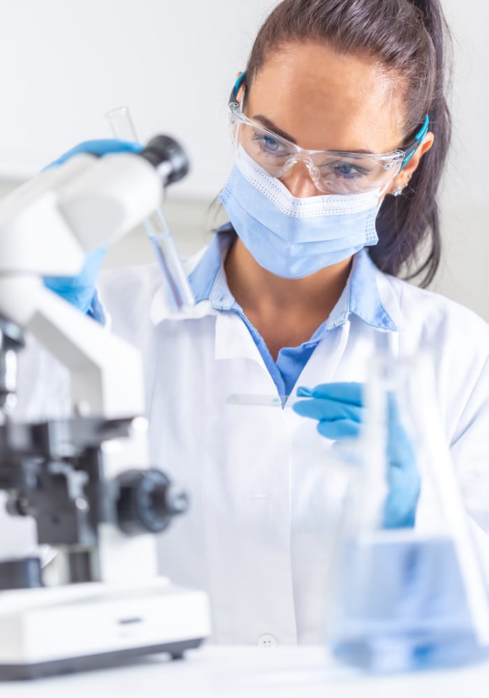 Female scientist wearing protective goggles and mask while examining samples under a microscope, ensuring safety and precision in laboratory testing.
